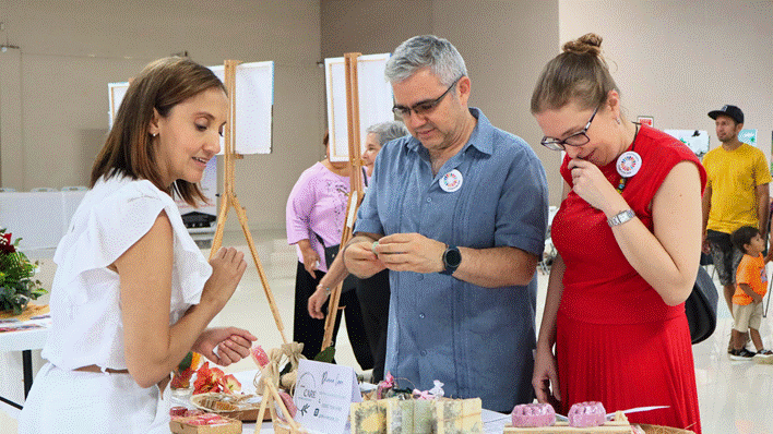 Krissia, emprendedora del sector de jabones, presenta sus productos a René Celaya, jefe de Misión de la Organización Internacional para las Migraciones (OIM), y a Gabriela Mata, coordinadora de Programas de ONU Mujeres, durante una Feria de Emprendedoras. Crédito: Mariana Álvarez.